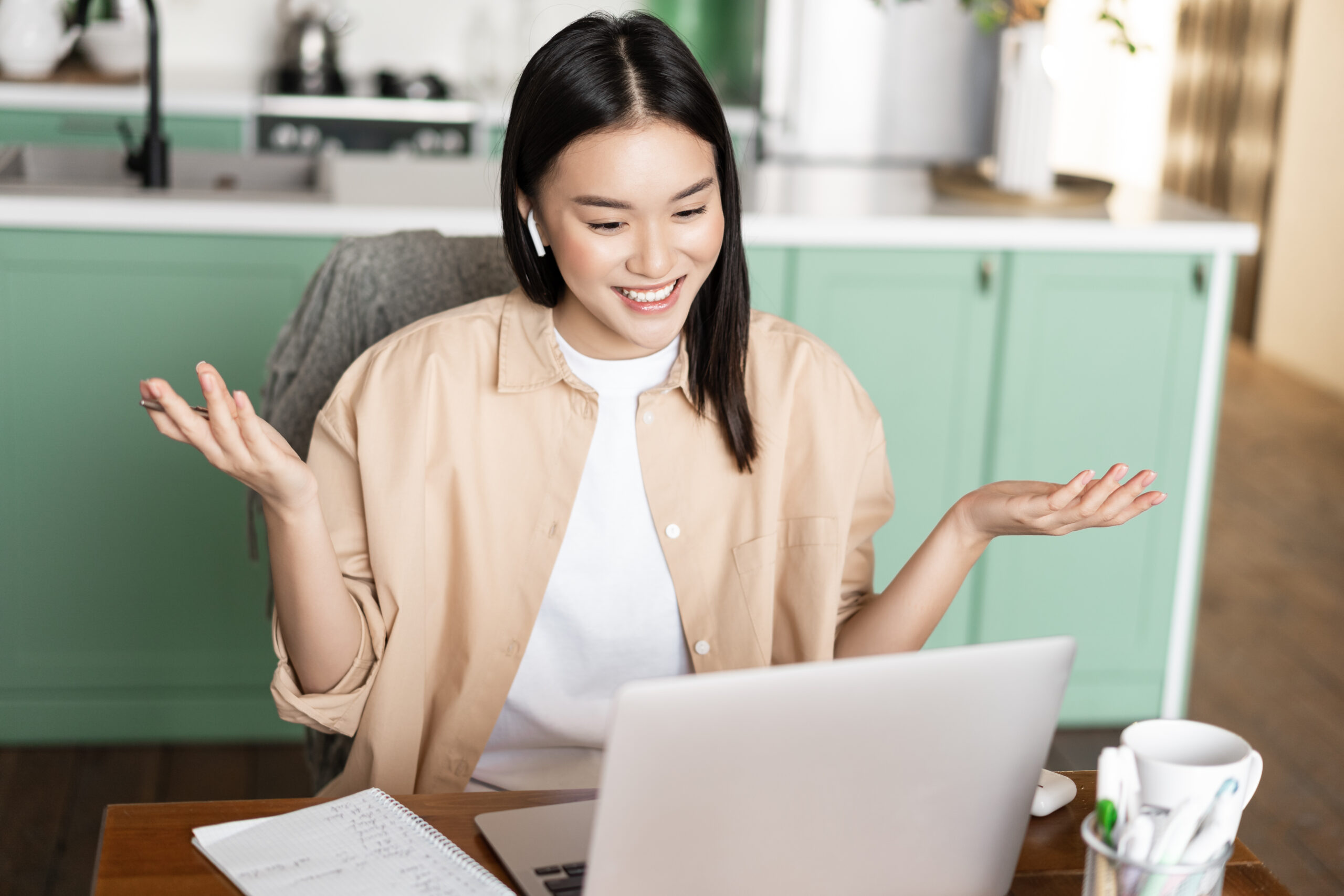 Asian woman tutor having online classes from home, working via laptop, having video call conference, wearing headphones, sitting at table surrounded by study material